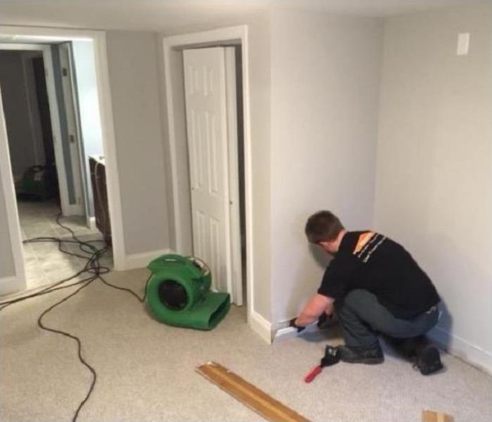 SERVPRO technician removing baseboard in flood damaged room. SERVPRO drying equipment in background