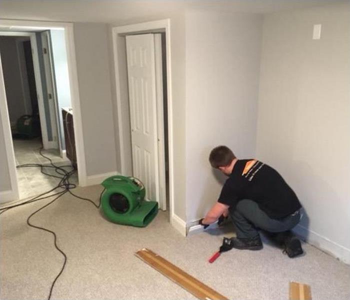 SERVPRO tech removing baseboard in water damaged room; SERVPRO drying equipment being used in room
